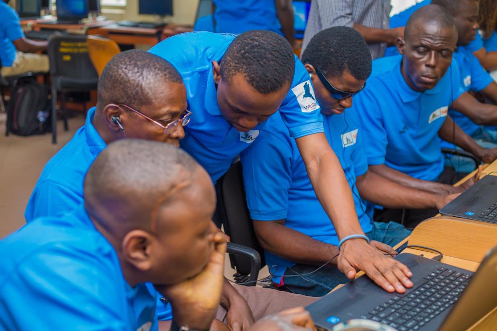 A man in a blue T-shirt with a laptop in front of him explains something to four other men who are all seated and listening to him.