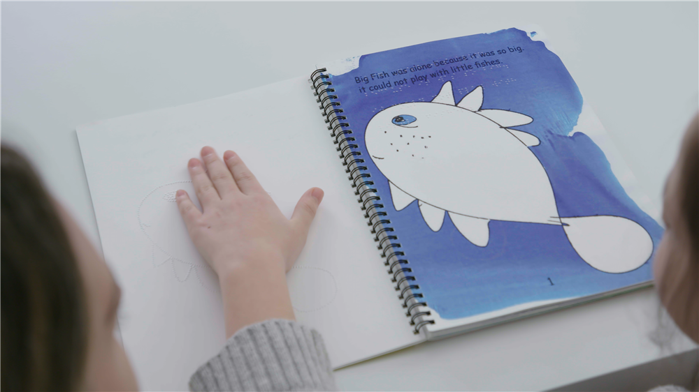 This image shows a close-up of an open book with a tactile picture that a child is exploring with their hand. The page visible contains a large printed fish and text that reads "Big Fish was alone because it was so big. It could not play with little fishes." The tactile image is a raised outline of the same fish, allowing a child, possibly with visual impairments, to feel and understand the picture through touch. The book appears to be an educational tool designed for inclusive learning, promoting accessibility and catering to the needs of children with different abilities. The image embodies themes of understanding, inclusivity, and the importance of providing equal opportunities for learning and engagement for all children.