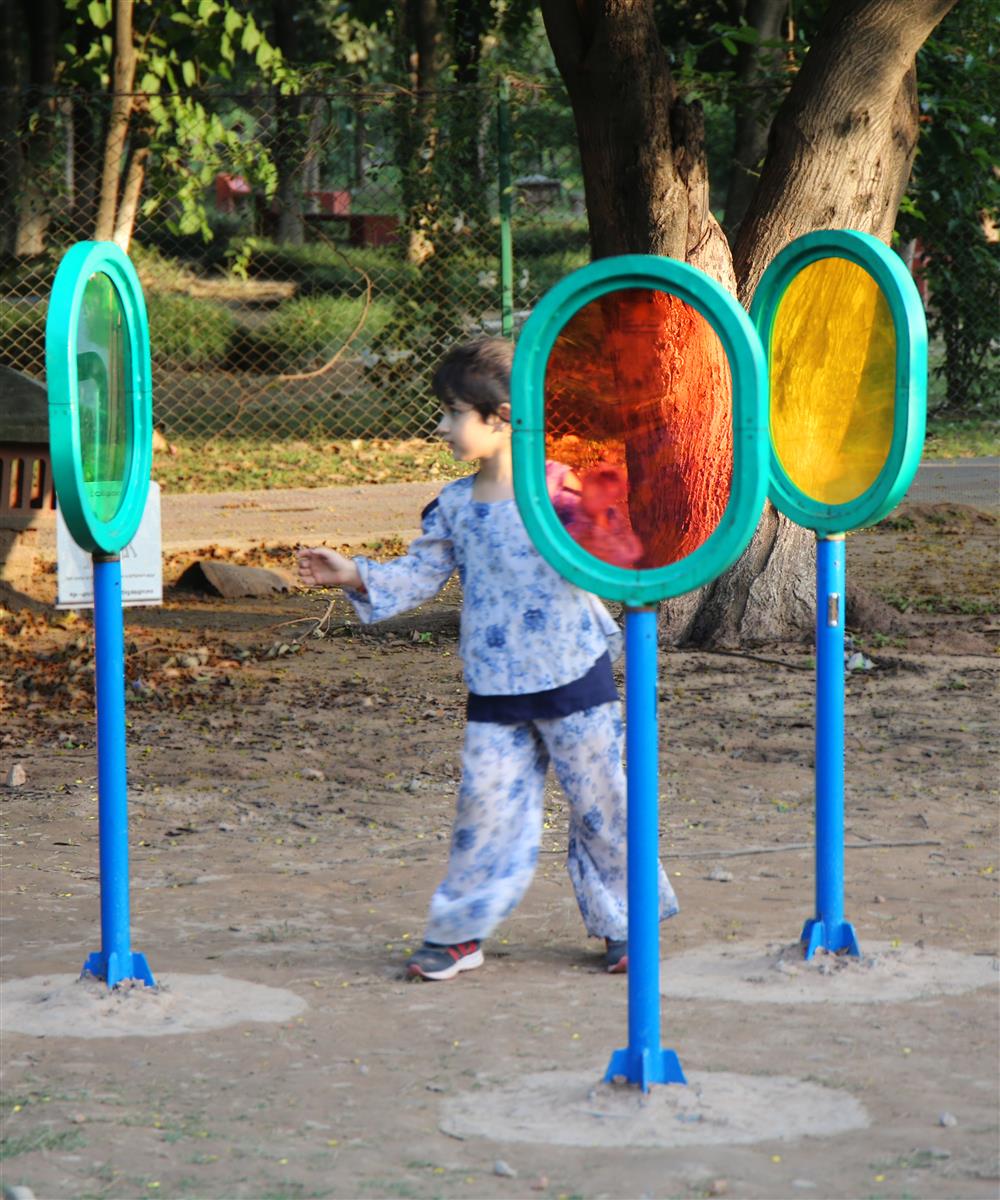 A small boy is standing in the center of three differently colored see through glass ovals, that are part of an accessbile playground, with a large tree in the background. 