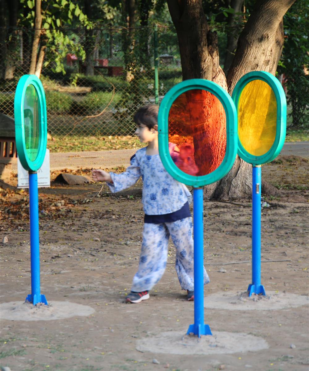 A small boy is standing in the center of three differently colored see through glass ovals, that are part of an accessbile playground, with a large tree in the background.  