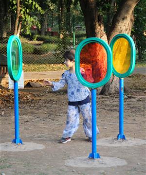 A small boy is standing in the center of three differently colored see through glass ovals, that are part of an accessbile playground, with a large tree in the background.  