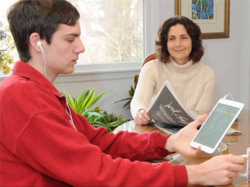 Young man wearing headphones reads highlighted text on a tablet, while sitting at a table with his mom who is reading the newspaper.