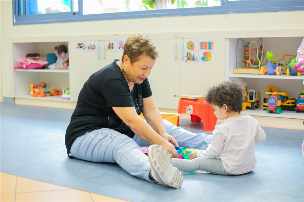 This image depicts a heartwarming scene of an older woman and a young child sitting on a soft mat on the floor, engaged in play. The woman, dressed casually in a black top and light blue jeans adorned with embellishments, is smiling gently as she interacts with the child. The toddler, wearing a white long-sleeve top and gray pants, is focused on colorful building blocks. The background suggests they are in a child-friendly environment, possibly a daycare or preschool, with toys and educational materials neatly stored and child artwork displayed. The interaction reflects a caring relationship, likely emphasizing the importance of intergenerational connections, nurturing, and learning.