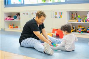 This image depicts a heartwarming scene of an older woman and a young child sitting on a soft mat on the floor, engaged in play. The woman, dressed casually in a black top and light blue jeans adorned with embellishments, is smiling gently as she interacts with the child. The toddler, wearing a white long-sleeve top and gray pants, is focused on colorful building blocks. The background suggests they are in a child-friendly environment, possibly a daycare or preschool, with toys and educational materials neatly stored and child artwork displayed. The interaction reflects a caring relationship, likely emphasizing the importance of intergenerational connections, nurturing, and learning.