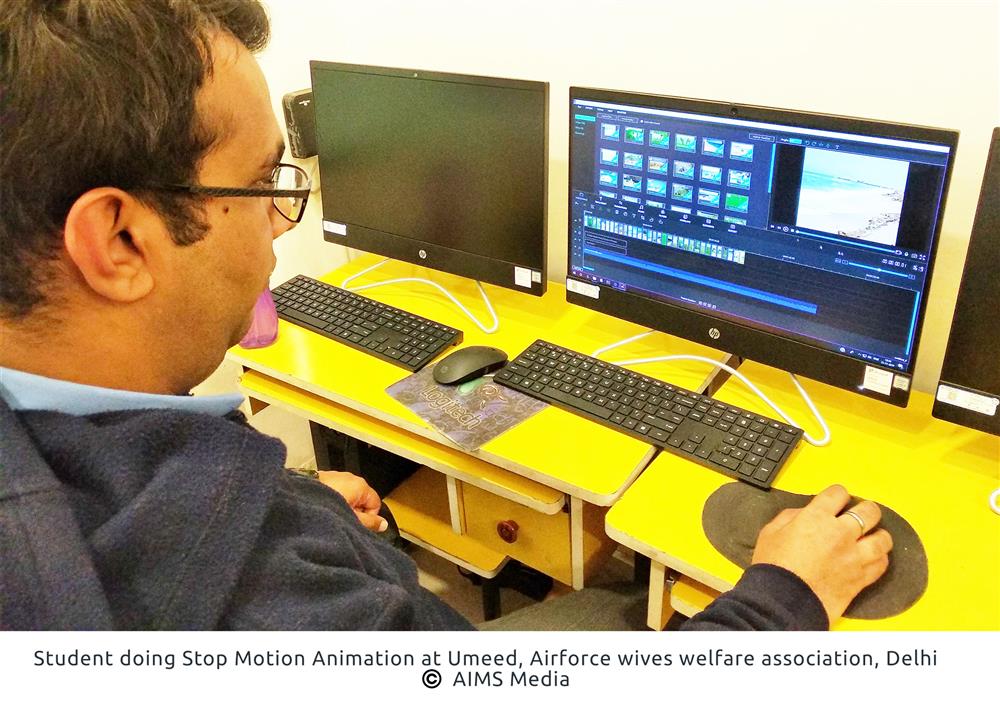 A male student is in front of a computer screen. He is holding a computer mouse in his right hand. On the screen are various forms and graphs. The mouse and keyboard rest on a yellow desk.