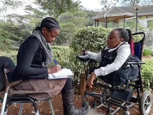 A woman in a wheelchair talks to another woman who has a crutch beside her chair. She is writing. They sit in a garden, and there are green plants behind them.