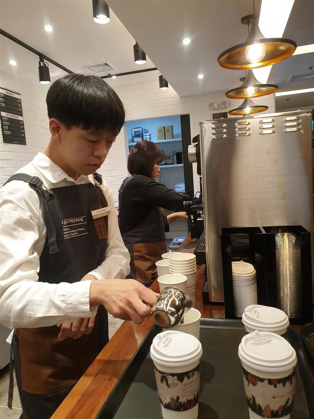 A young man wearing an apron stands behind a coffee machine in a coffee shop. He is pouring milk into a takeaway coffee cup. A woman in an apron is making coffee at a second machine in the background.