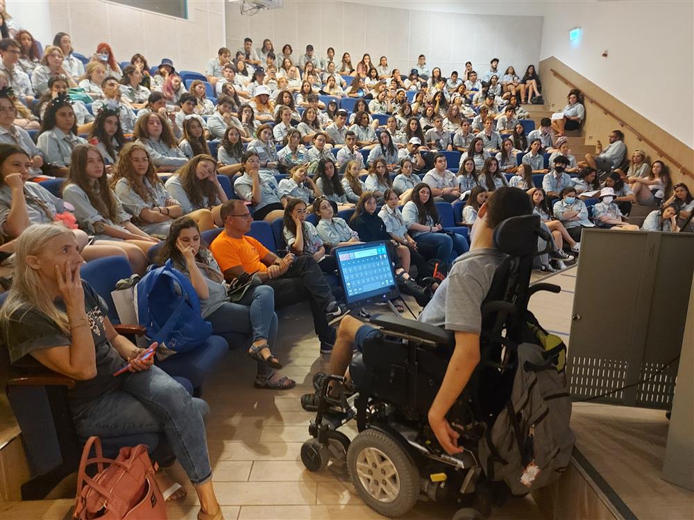 A man in an electronic wheel chair with an electonic tablet attached on the armrest is speaking in an auditorium filled with students in uniform.