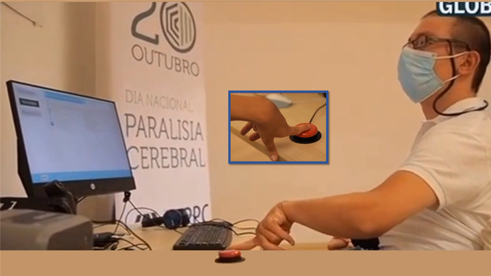 A man who appears to have cerebral palsy tries to press a button with his index finger and a computer monitor and keyboard in front of him. A white tarpaulin beside him is printed with the text "20 Outubro Dia Nacional Paralisia Cerebral".