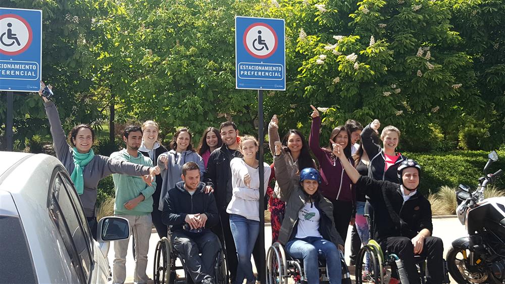 A group of young adults, with and without physical disabilities standing in front of a parking space for people with wheelchairs.