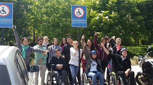 A group of young adults, with and without physical disabilities standing in front of a parking space for people with wheelchairs.
