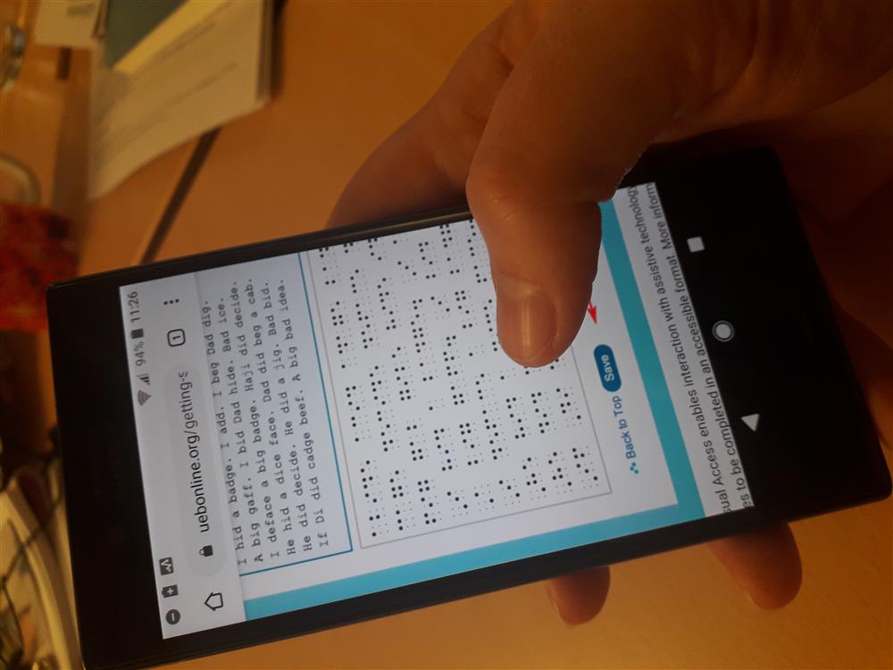 Hand holds a mobile phone, while scrolling through a Braille online learning tool.