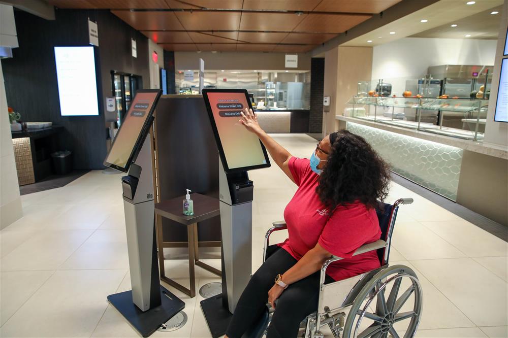 A woman in a wheelchair is ordering food on one of two touchscreen devices that are positioned in the center of Eli Lilly's accessible and modern cafeteria. 