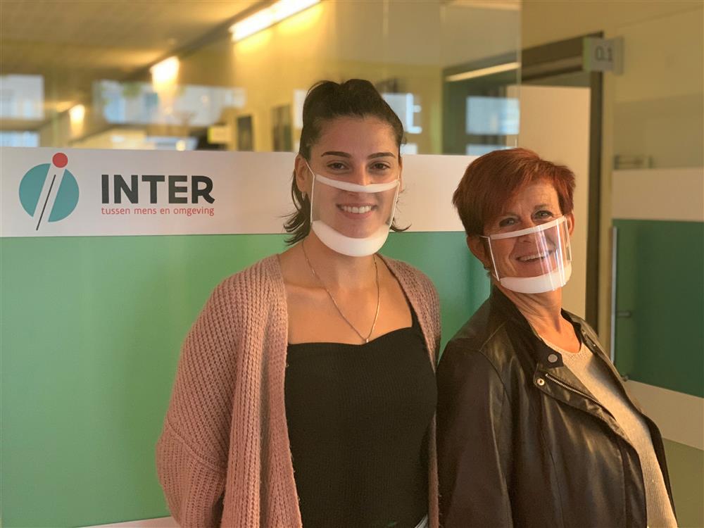 An office setting, in which two women wearing transparent face masks, stand in front of a white and green wall with a lettering saying  "Inter. between people and environment" in dutch. 