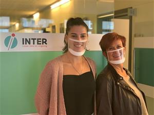 An office setting, in which two women wearing transparent face masks, stand in front of a white and green wall with a lettering saying "Inter. between people and environment" in dutch. 