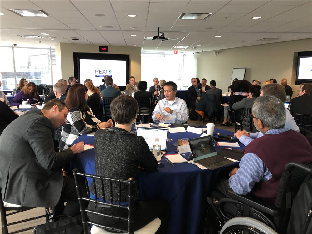 There are five round tables in a conference room. Six people sit around each table with laptops and papers in front of them. A presentation is being projected on a screen at the front of the room.