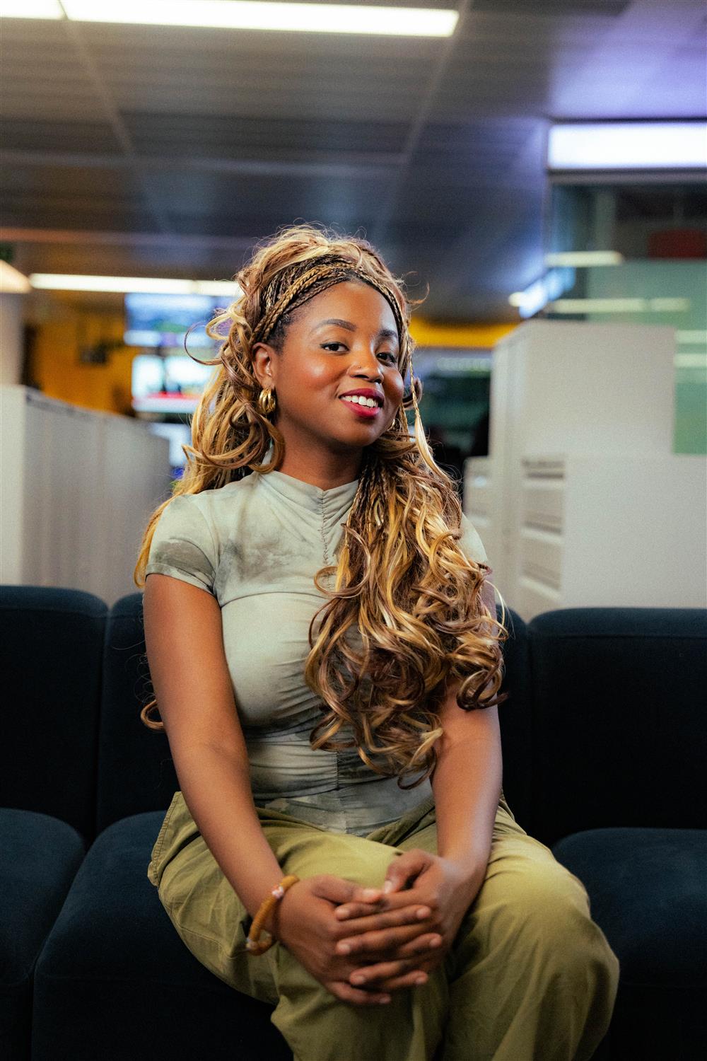 A young African woman with braided hair sits confidently in a modern office. Her expression is poised, reflecting professionalism and empowerment. The setting suggests she might be in a media or corporate environment, symbolizing diversity and representation in the workplace.