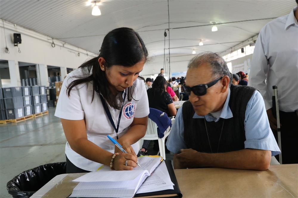 A man and a woman are at a desk with paperwork in front of them. The man is seated on the right of the picture wearing dark glasses. The woman is standing next to him and writing on a form.