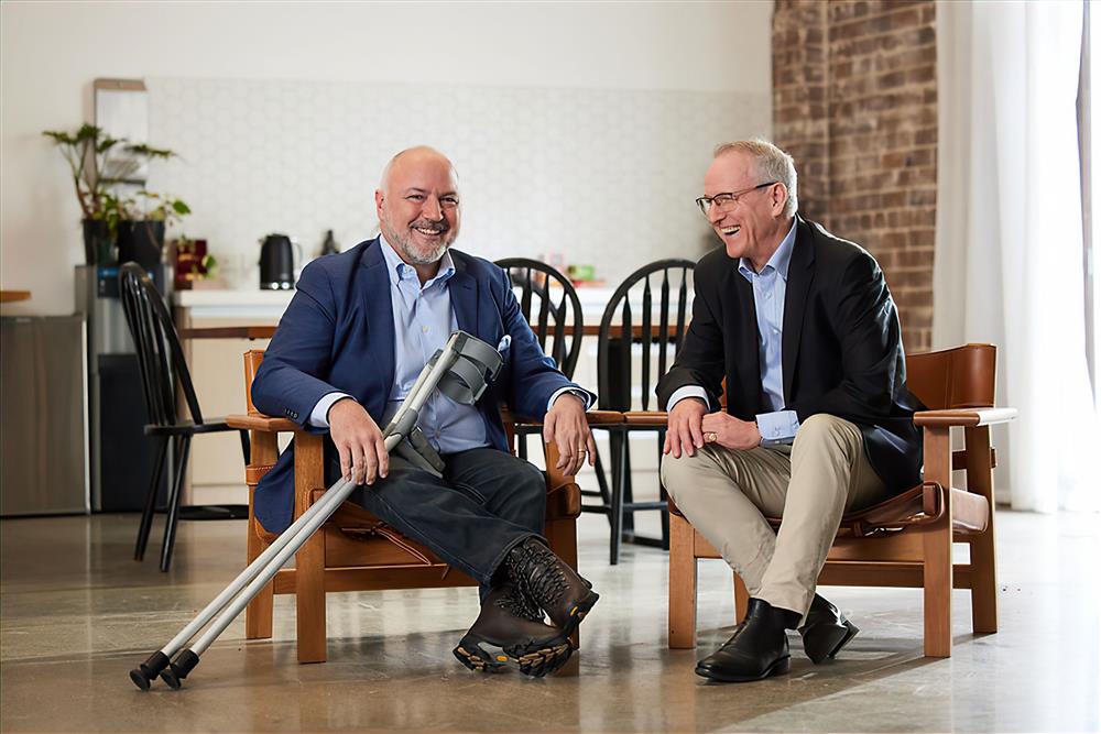 The photo depicts two men sitting in chairs facing each other with warm, friendly expressions. The man on the left has a prosthetic leg, which is clearly visible, and he is holding crutches. The environment suggests a casual, possibly professional setting. Both men are dressed in business casual attire. The presence of the prosthetic leg may subtly suggest themes of overcoming adversity, resilience, and normalizing disability in everyday contexts, promoting a message of inclusivity and equality.