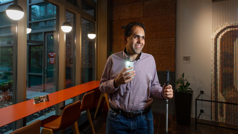 A blind man holding a white cane in holding up a smartphone and smiling into the camera.