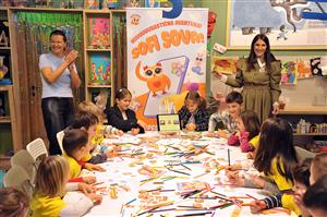 Children gather around a large table covered in colorful pencils and drawings, guided by two smiling educators. The cheerful setting promotes creativity and learning through play, emphasizing equal opportunities for children to express themselves and collaborate joyfully.