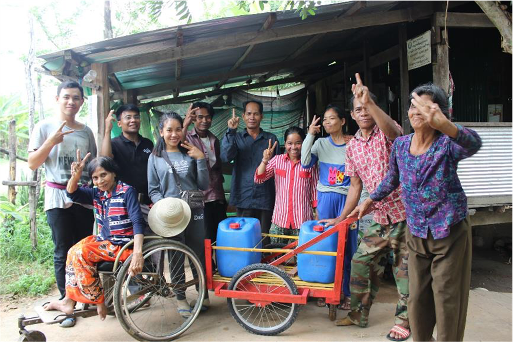A group of 5 men women, one sitting in a wheelchair with a transportation device attached, stand in front of a wooden shed each holding up the "peace sign" with their hands. 
