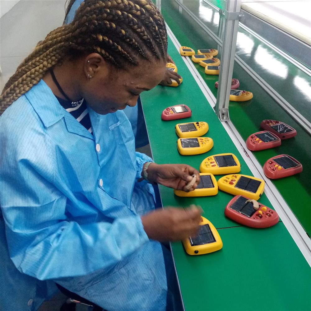 An African woman in a work uniform meticulously assembles small solar-powered devices on a production line. Her focused expression and the bright colors of the devices symbolize the empowerment of women in technology and sustainable energy industries.