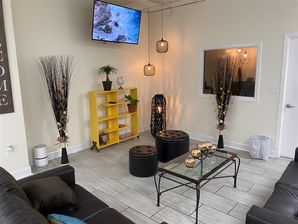 A living room with two couches perpendicularly placed across a hanging lamp and a television mounted on the wall with a shelf below. Beside the door is a painting. Candles on top of a glass center table in the center of the room.