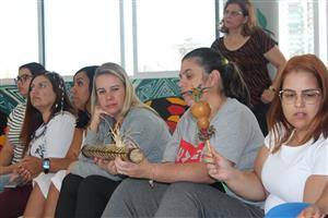 The photo shows a group of women engaged in an activity that appears to be cultural or spiritual in nature. One woman in the foreground is holding a traditional object, which could be used in indigenous rituals or ceremonies. The participants seem focused and reflective, suggesting a moment of shared experience or learning. The setting includes a mix of casual and somewhat traditional attire, indicating a diverse gathering. The presence of cultural elements and the attentive atmosphere reflect themes of respect, community, and cultural appreciation.