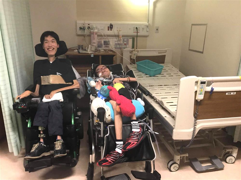 Two men appearing to have different severity of muscular dystrophy seated in their electronic wheelchair inside a hospital room with a hospital bed on the right side of the photo and a light source behind them.