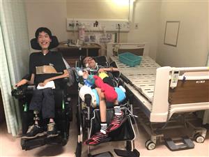 Two men appearing to have different severity of muscular dystrophy seated in their electronic wheelchair inside a hospital room with a hospital bed on the right side of the photo and a light source behind them.