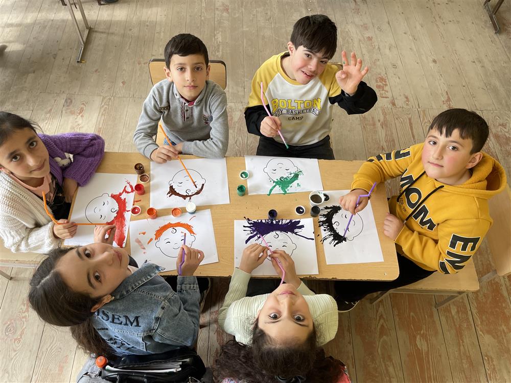 The photo shows a group of six children engaging in an art activity. They are seated around a table, each with a piece of paper in front of them, painting or drawing faces with various expressions. The children appear focused and happy, with some looking up and smiling at the camera. One child is making a friendly wave. Their diverse appearances reflect a natural and harmonious blend of backgrounds, emphasizing inclusivity. The environment seems to be a classroom, which is bright and welcoming. The presence of paint pots and brushes suggests a creative learning atmosphere.