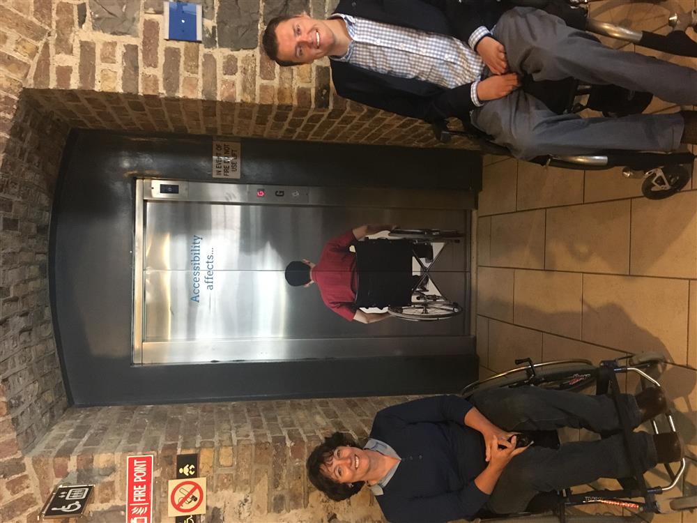 Two people on wheelchairs in front of an elevator that has a picture of person with a wheelchair stuck on the doors saying 