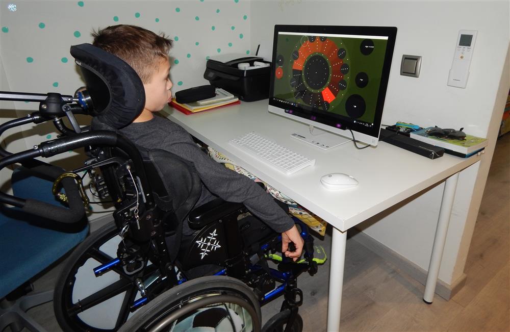 A young boy sitting in his wheelchair in front of a screen on a white table. He ist playing the eyeharp, a digital music instrument that can be controlled with eye or head movements. 