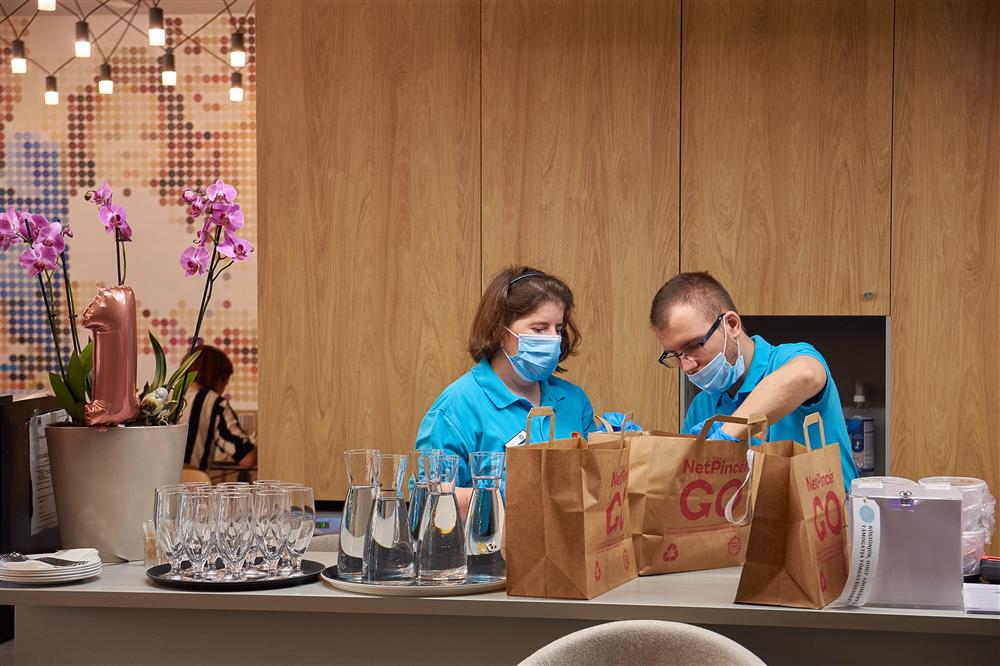 A man and a woman wearing hygiene masks pack food in paper bags, which sit on a counter in front of them. On the counter are also flowers and full carafes of water and empty water glasses.