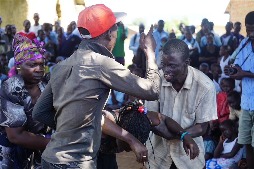 A scene in a public square in Africa, with two men and one woman steady one women who is unconscious or has fallen. All have serene facial expressions, one of the man is talking intensily to a crowd of people observing and taking pictures. 
