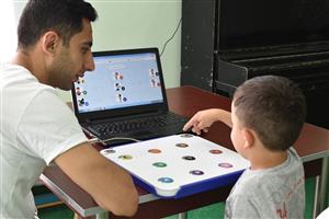 A boy points to the keyboard with different colored buttons connected to the laptop while the man seated beside him is watching over the boy.