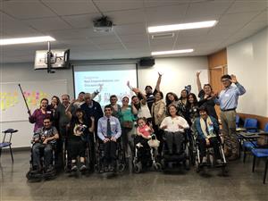 A group of Chilean persons appearing to have different motor disability compressed for a photo with hands raised in the air in front of a beamer screen that reads "Programa Red Emprendedores con Discapacidad".