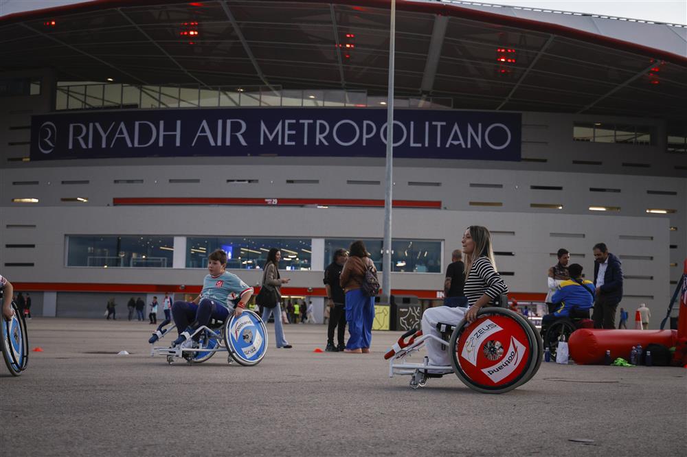 The photo shows a woman and a child using sports wheelchairs in an open plaza outside the Riyadh Air Metropolitano stadium. They participate confidently in an inclusive activity alongside others nearby.
