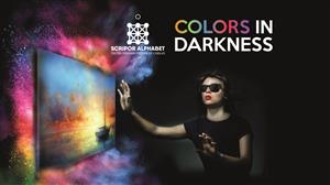 A woman wearing sunglasses with hands raised up trying to touch a painting on a wall. On the upper right side of the photo, the phrase "Colors in Darkness" written beside the Scripor Alphabet logo.