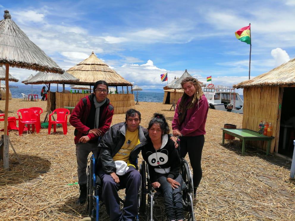 A man and woman sitting in their wheelchairs and a man and woman standing behind them in a beachfront with Cabanas around.