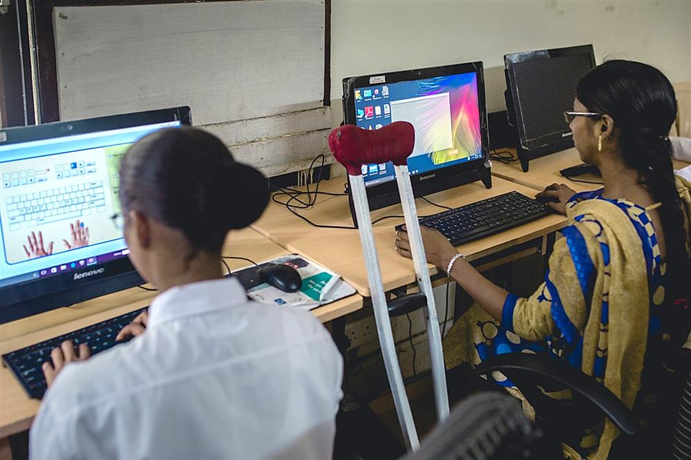 Two young women sit next to each other in front of two computers, with a crutch leaning on the desk between them. One woman is doing a typing exercise.