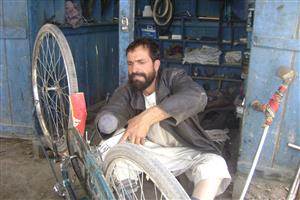 A man who has had his right hand and left leg amputated sits next to an upturned bike in a workshop and examines the chain.