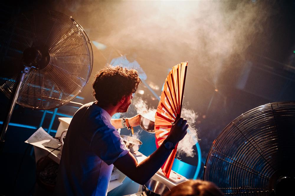 A man performs creatively with large fans, mist, and dramatic lighting, blending science and art. His expressive gestures and concentration highlight how artistic expression unites people across backgrounds and abilities.