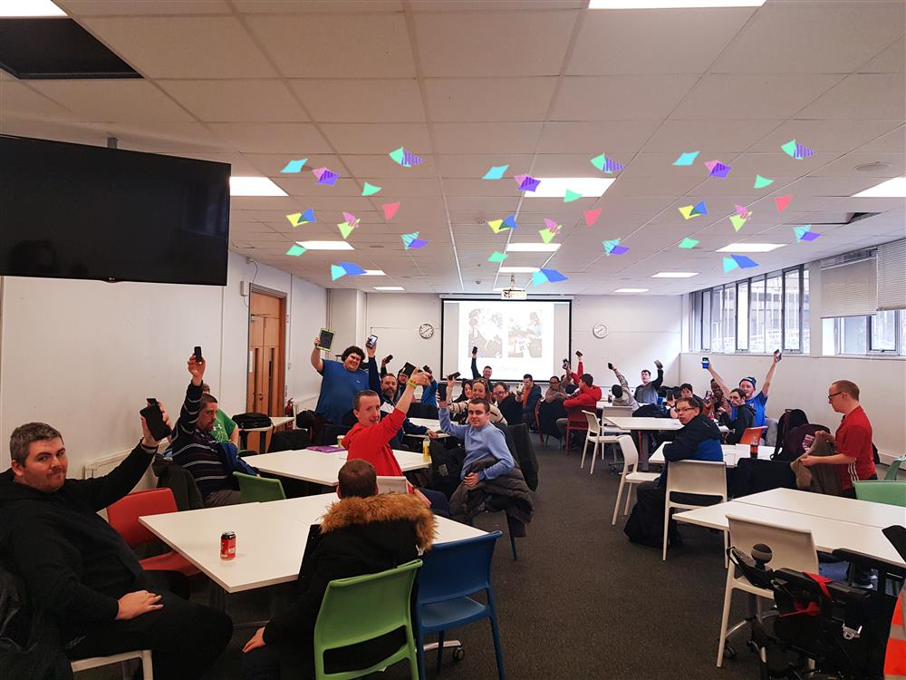 A mixed group of men in different ages and appearing to be in the autism spectrum seated in groups scattered in a hall raising one of their hands holding a mobile phone.