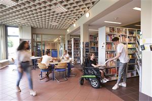 This image shows a bright and inviting library space with individuals engaged in various activities. In the foreground, a person in a motorized wheelchair is interacting with a standing person, suggesting an atmosphere of inclusivity and assistance. In the background, two individuals are playing chess at a table, while another person walks by, blurred by motion, adding a dynamic feel to the scene. The library shelves are well-stocked, indicating a place of learning and resourcefulness. The environment promotes equality and tolerance, as people of different abilities and interests share the space harmoniously.