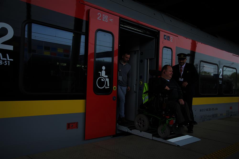 We are on a platform of a train station. One of the train conductors is standing by as a man with an electric wheelchair calmly exits the train over a long silver ramp, followed by another passenger. 