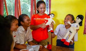 Two Indonesian students in school uniform giggling together with another student and a woman each holding a doll.
