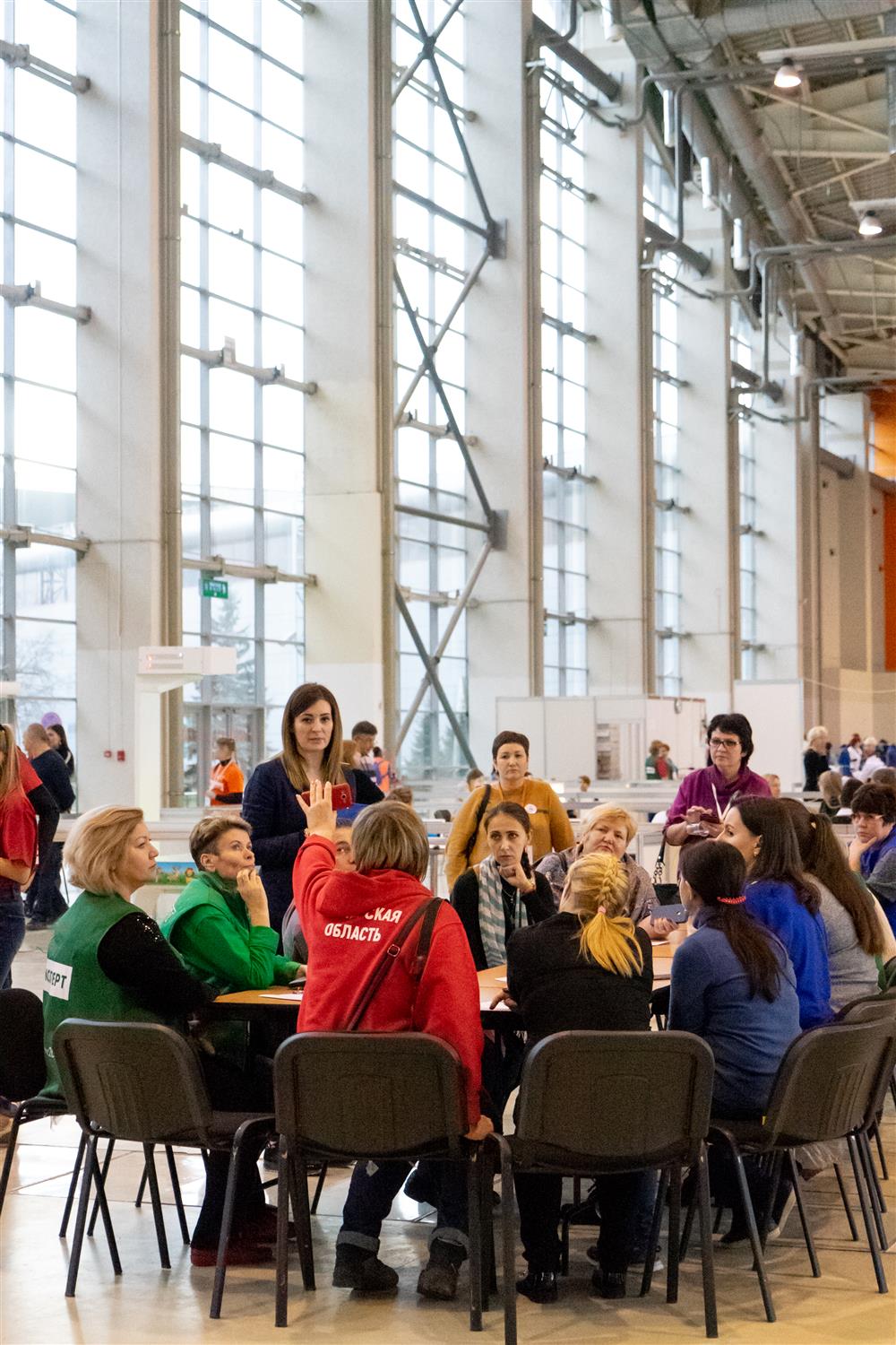 A total of ten female and male students sit at a round table. One of them is raising his hand. Several women stand behind the chairs. There is a tall class wall in the background.