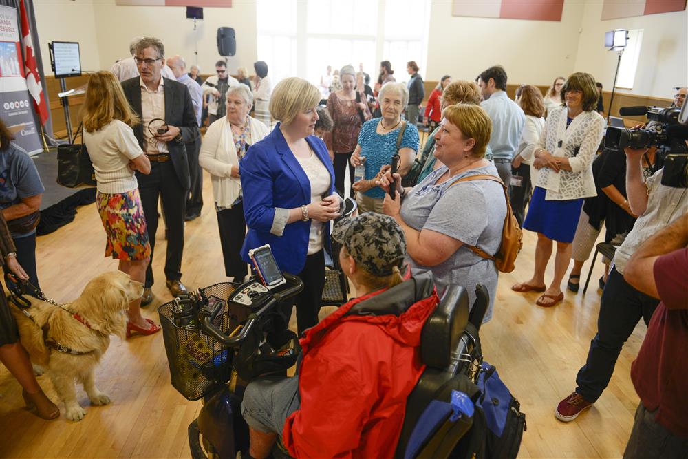 This image shows a bright conference room filled with persons with and without disabilites. In the foreground there are two women, one in an electric wheelchair and one blind, talking to a female state official.  